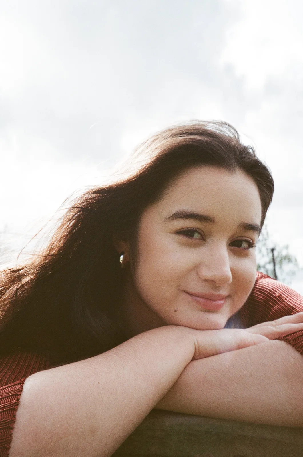 Clarissa Mae smiling outdoors, resting her chin on folded arms in a rust-coloured cardigan.