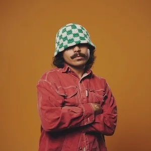 Sein Macey standing with folded arms in a red denim outfit and a green-and-white crocheted bucket hat against an ochre background.